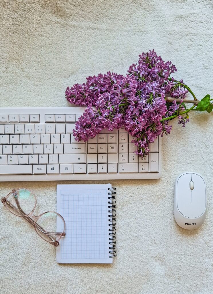 Flat lay of purple lilacs with a keyboard, glasses, and notebook. Ideal for work-from-home concepts.
